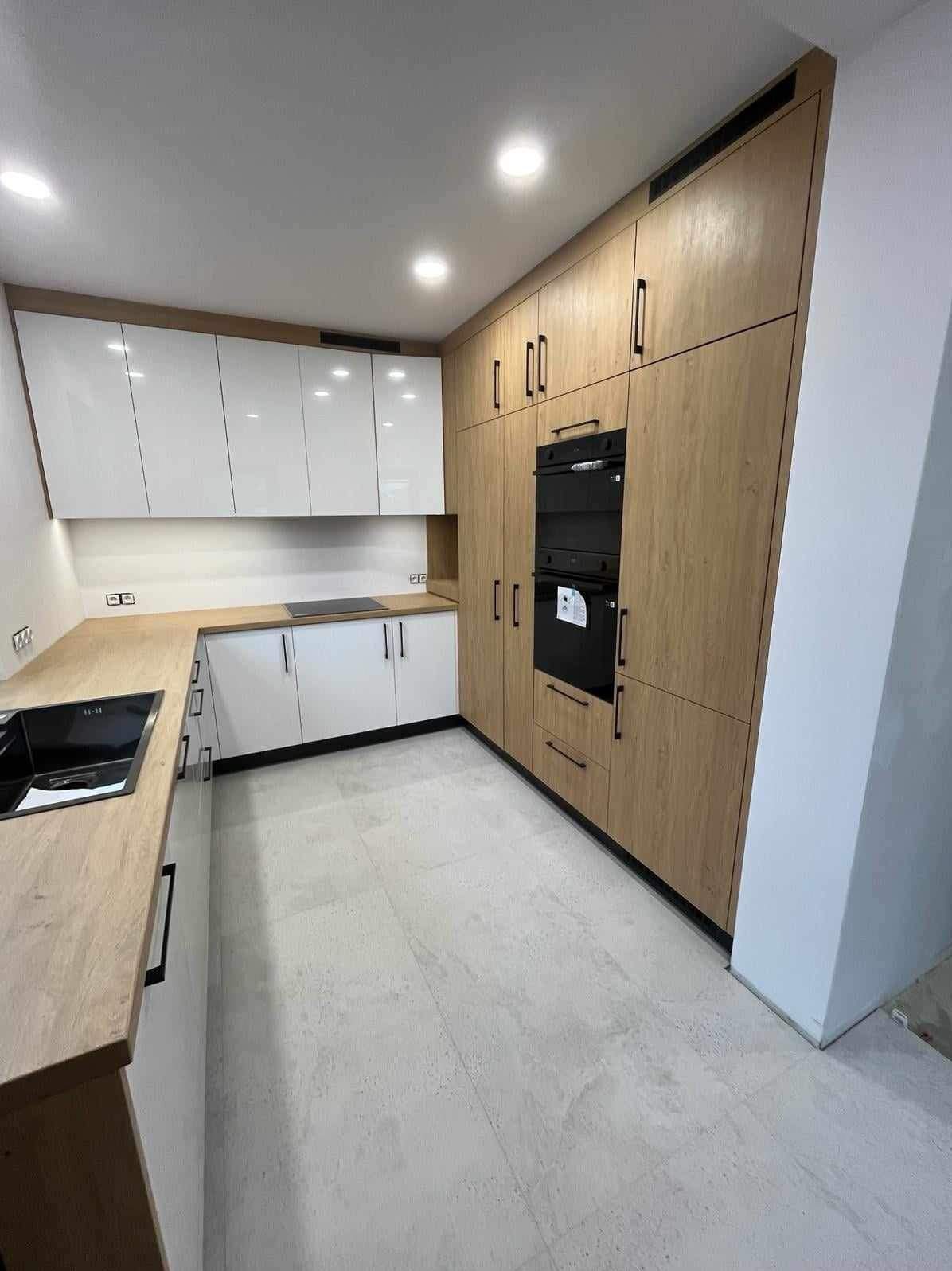 Nowoczesna kuchnia z białymi szafkami górnymi i dolnymi, drewnopodobnymi wysokimi szafami z czarnym piekarnikiem i czarnymi uchwytami, jasna podłoga.