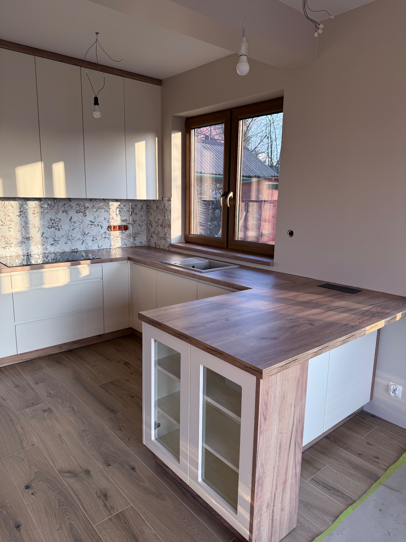 Jasna kuchnia z drewnianym blatem i wzorzystą ścianą nad blatem w Barwałdzie Dolnym. Widoczne szklane fronty szafek i drewniana podłoga. Oświetlenie w trakcie montażu.