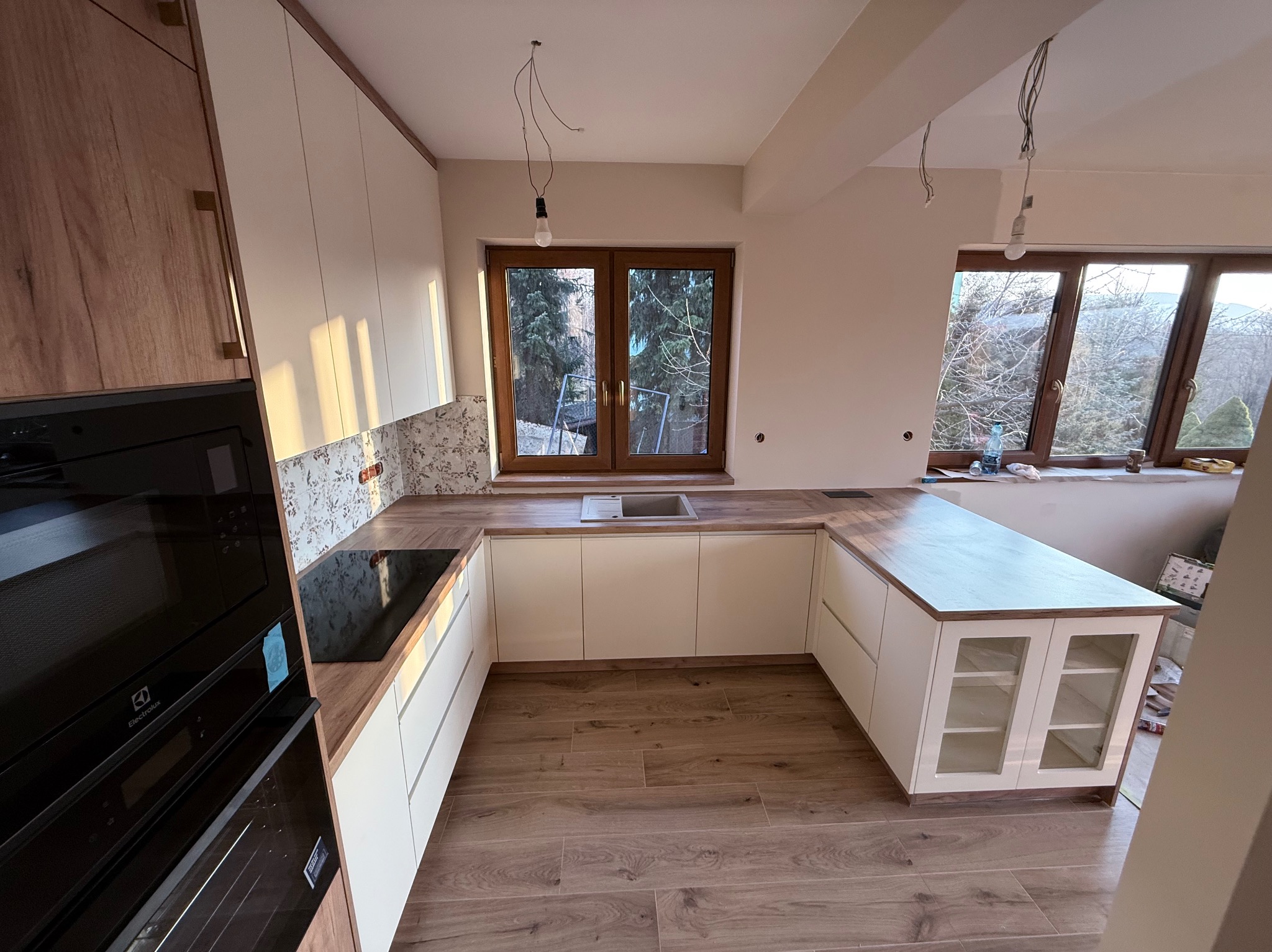 Jasna kuchnia na wymiar w kształcie litery U z drewnianym blatem i białymi frontami, widok na okno z zielenią. Nowoczesny design, jeszcze bez lamp.