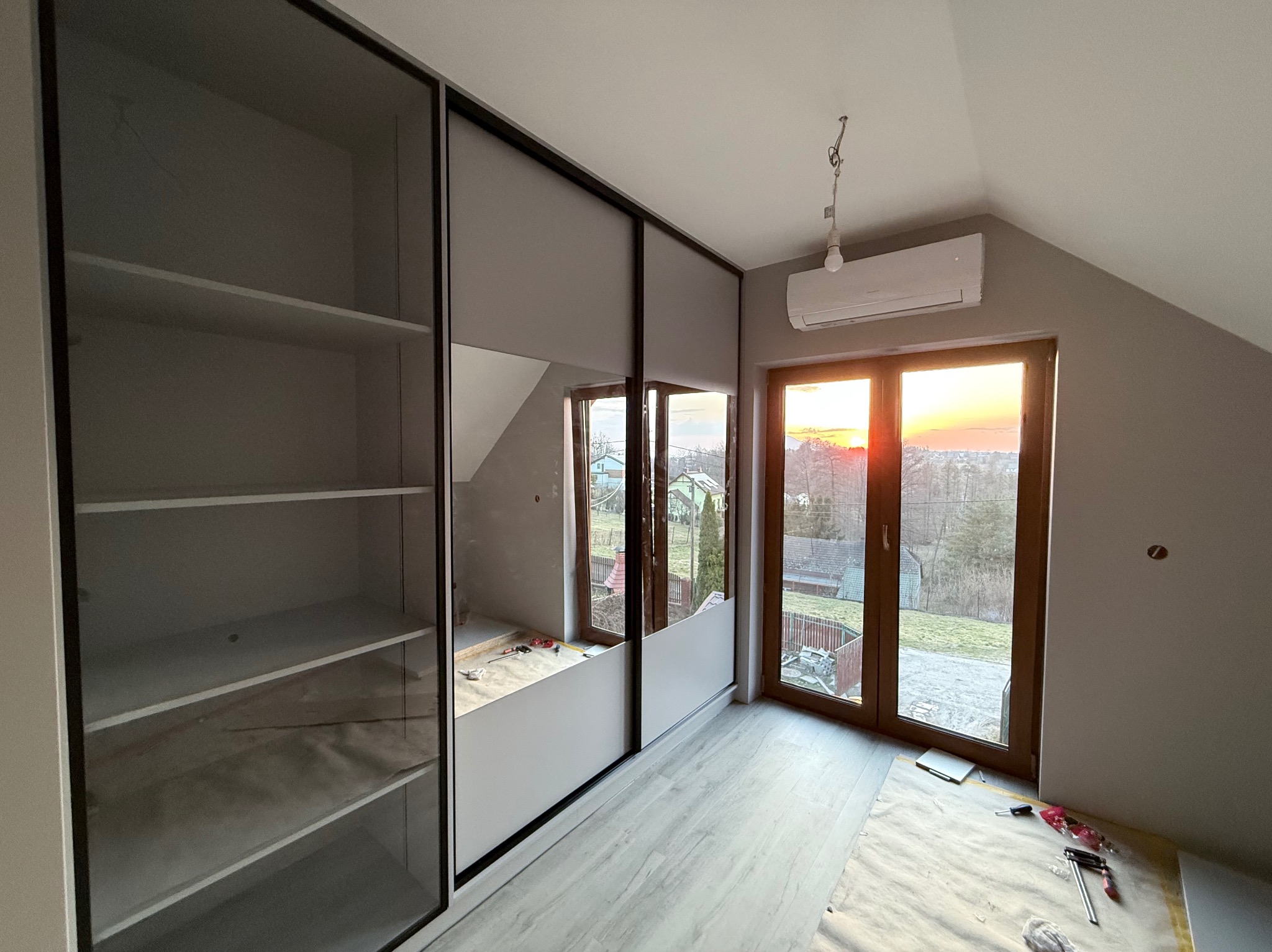 Szafa wnękowa z przesuwnymi drzwiami i lustrem, w jasnym pokoju na poddaszu. Widoczne narzędzia na podłodze sugerują montaż. Za oknem zachód słońca.