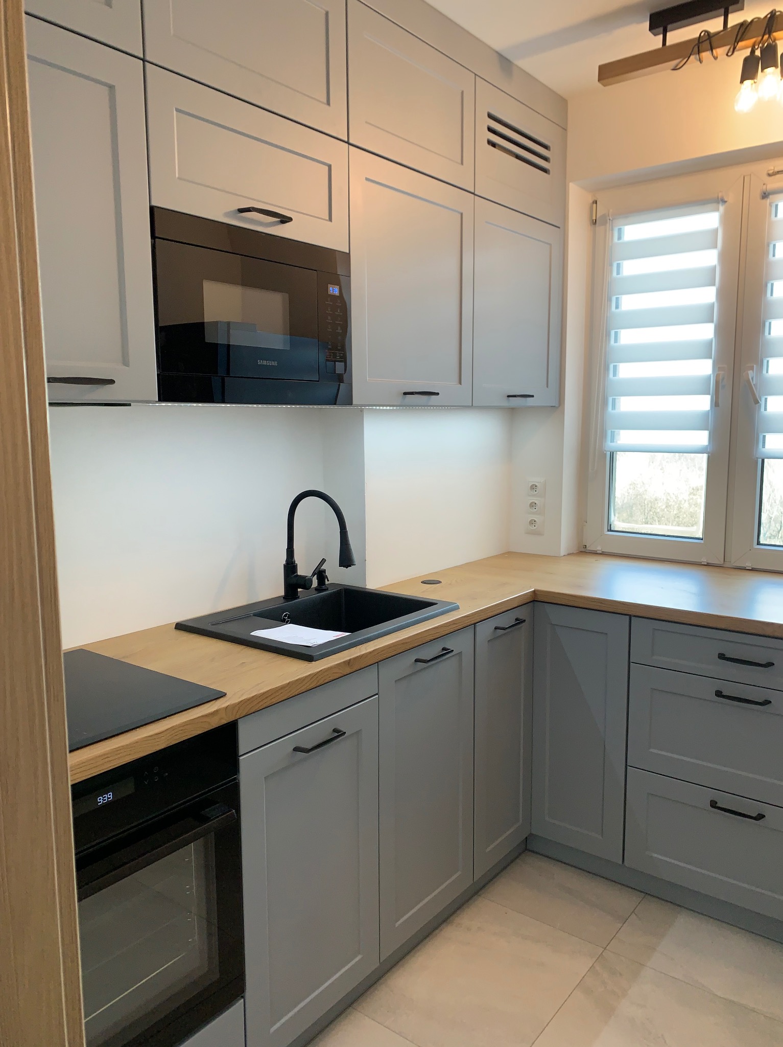 Szara kuchnia z drewnianym blatem i czarną armaturą. Widoczny piekarnik, mikrofalówka i okno z żaluzjami. Nowoczesny design, minimalistyczne uchwyty.