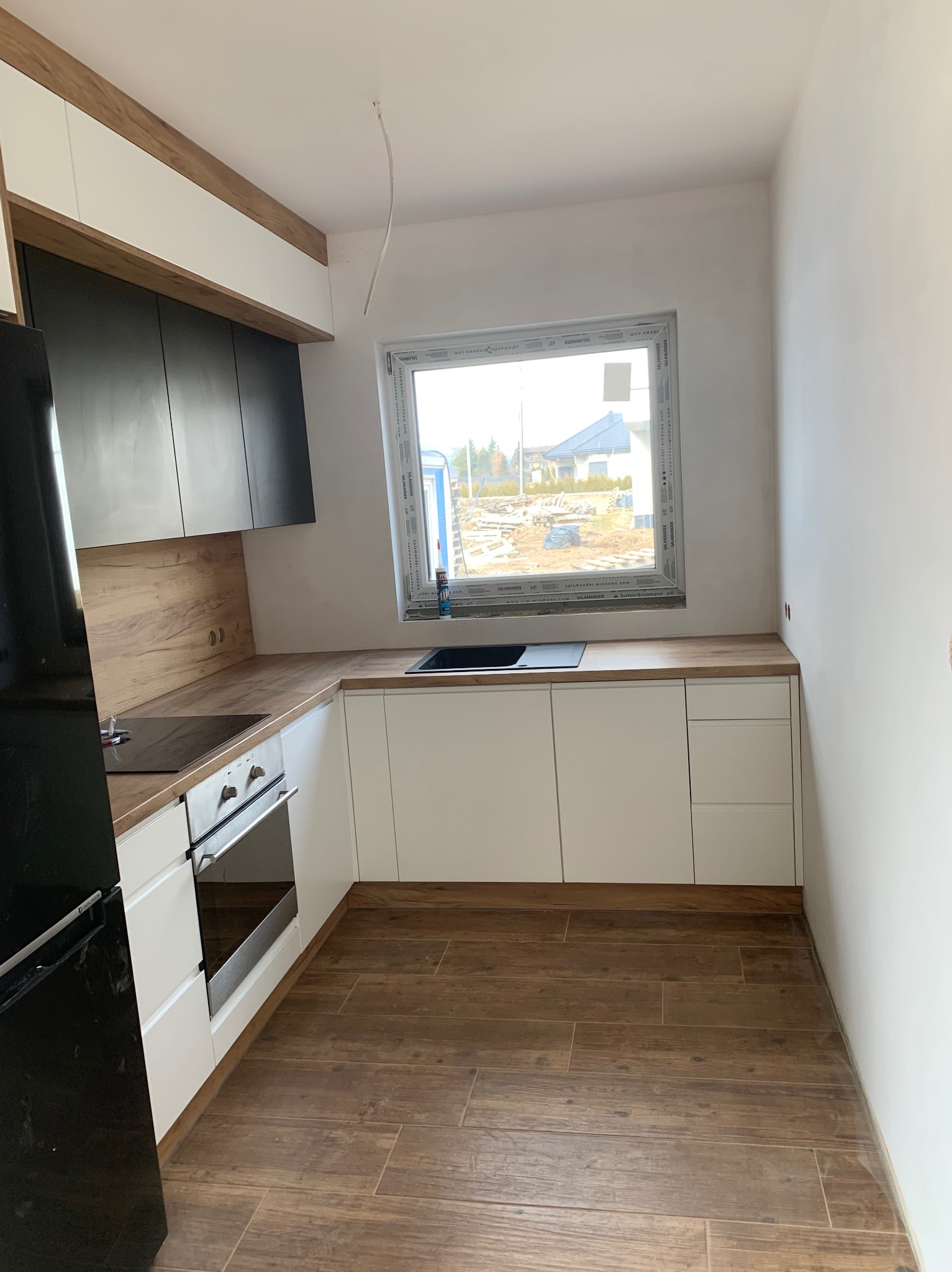Jasna kuchnia z drewnianymi akcentami: białe szafki dolne, czarne górne, drewniany blat i podłoga. Okno z widokiem na budowę. Minimalistyczny design.