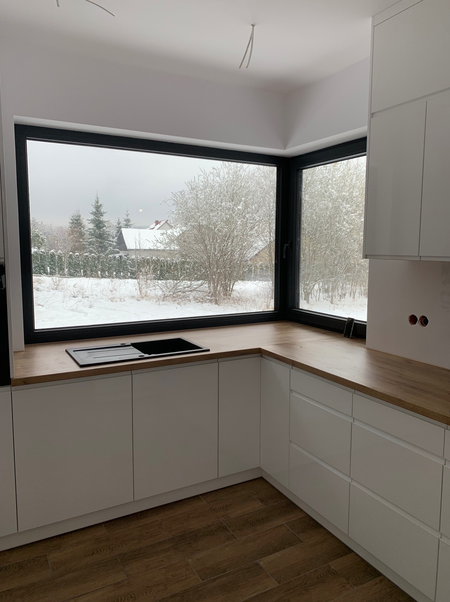 Nowoczesna kuchnia z białymi szafkami i drewnianym blatem, narożne okno z widokiem na zimowy krajobraz. Minimalistyczny design, czarny zlew.