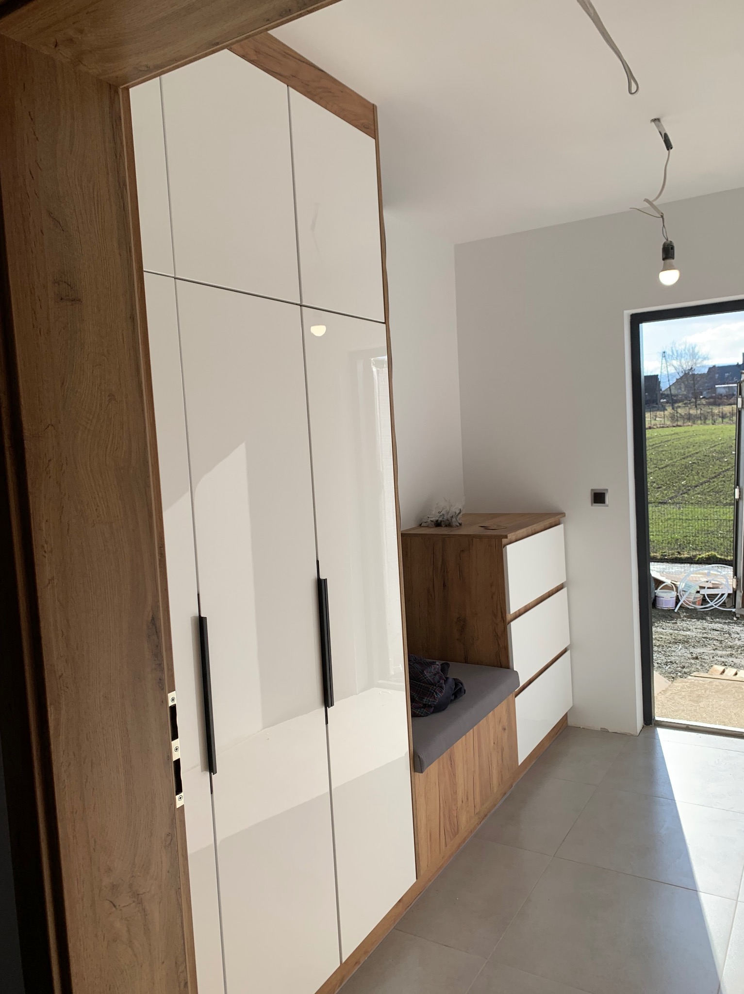 Nowoczesna szafa wnękowa z białymi frontami i drewnianym korpusem, połączona z siedziskiem i komodą w przedpokoju. Widok na zewnątrz przez przeszklone drzwi.