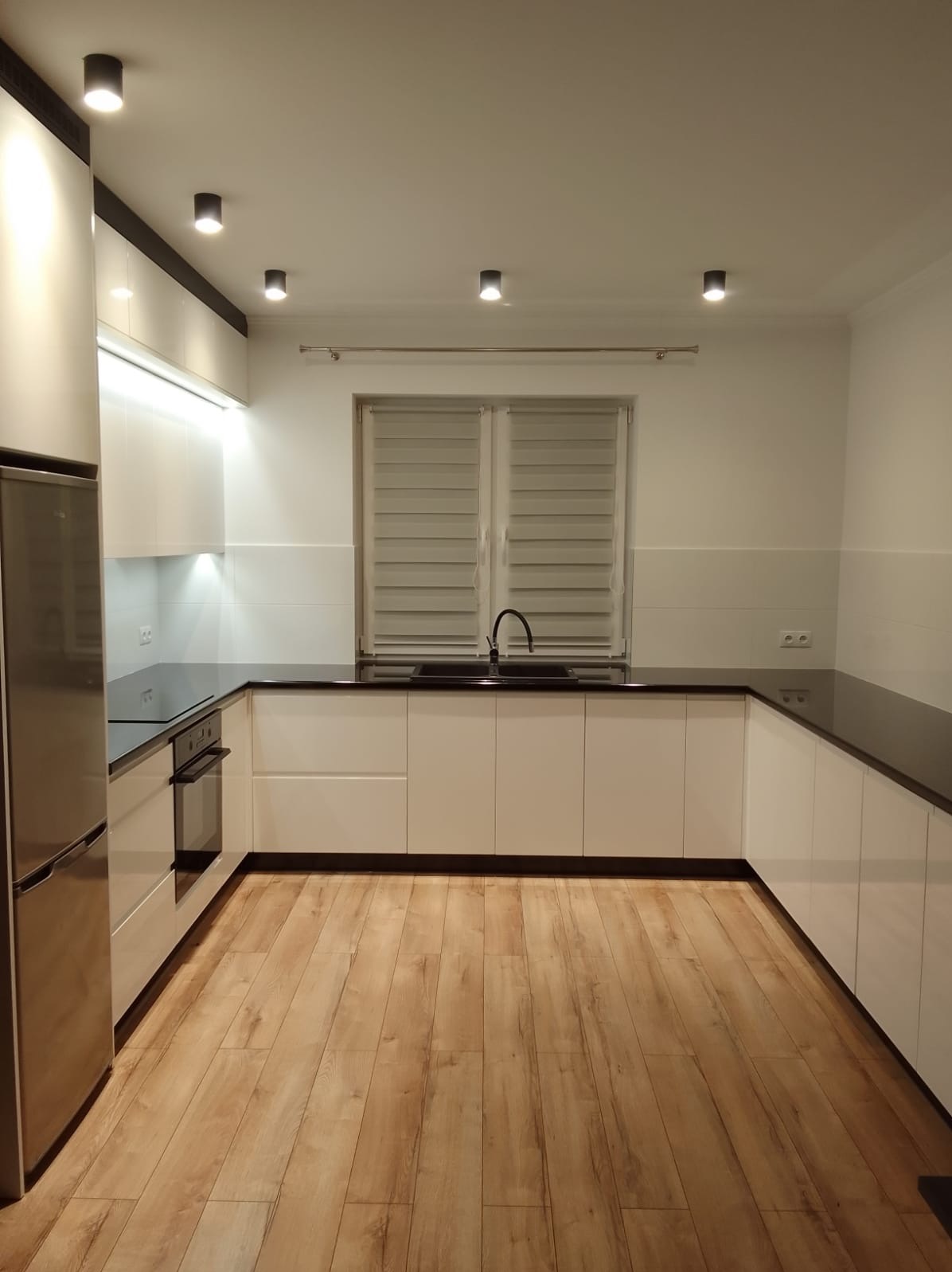 Nowoczesna, jasna kuchnia z białymi szafkami i czarnym blatem. Widoczne oświetlenie punktowe i drewniana podłoga. Minimalistyczny design.