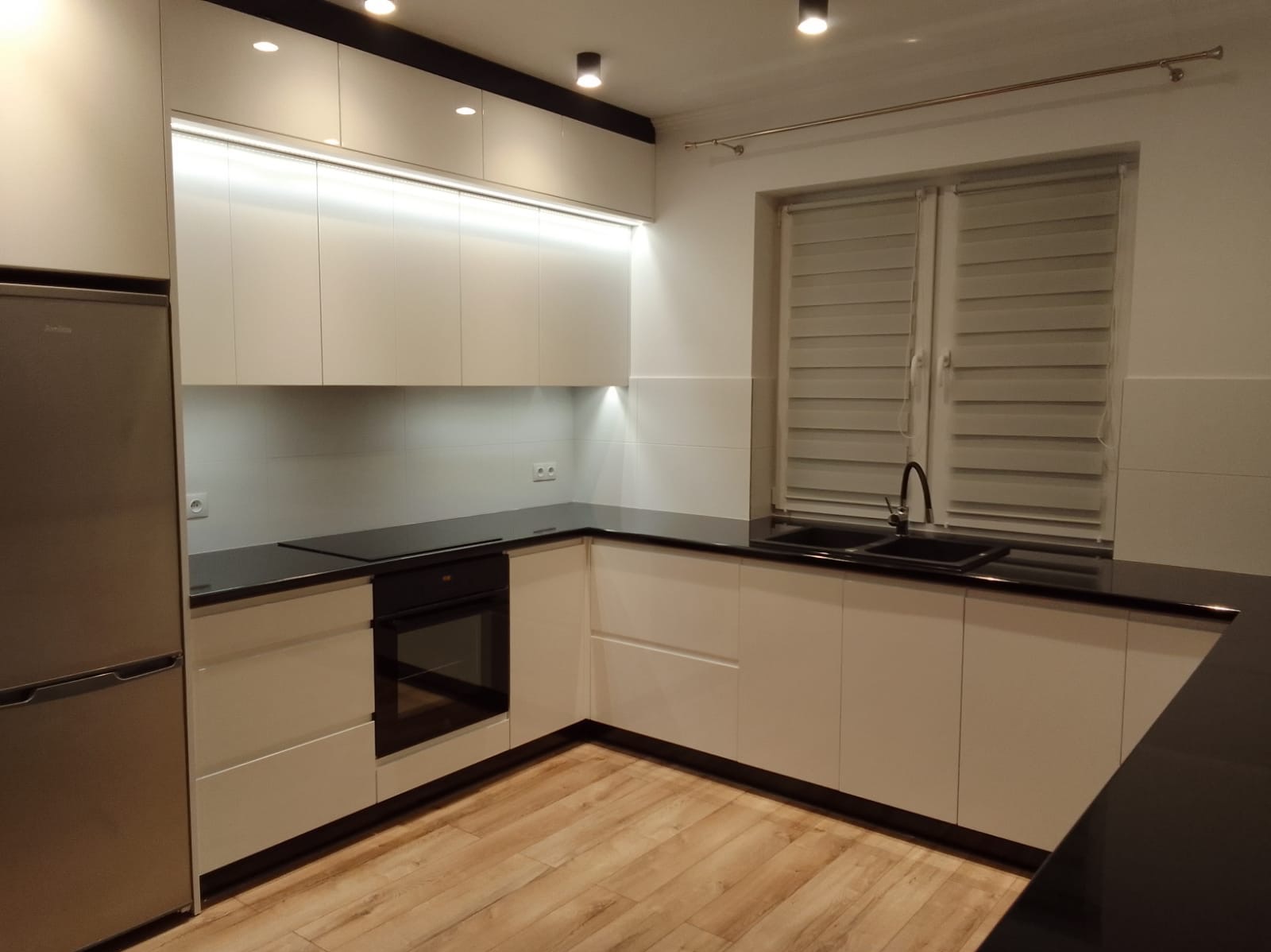 Nowoczesna kuchnia z białymi frontami i czarnym blatem. Widoczne oświetlenie podszafkowe LED i drewnopodobna podłoga. Minimalistyczny design.