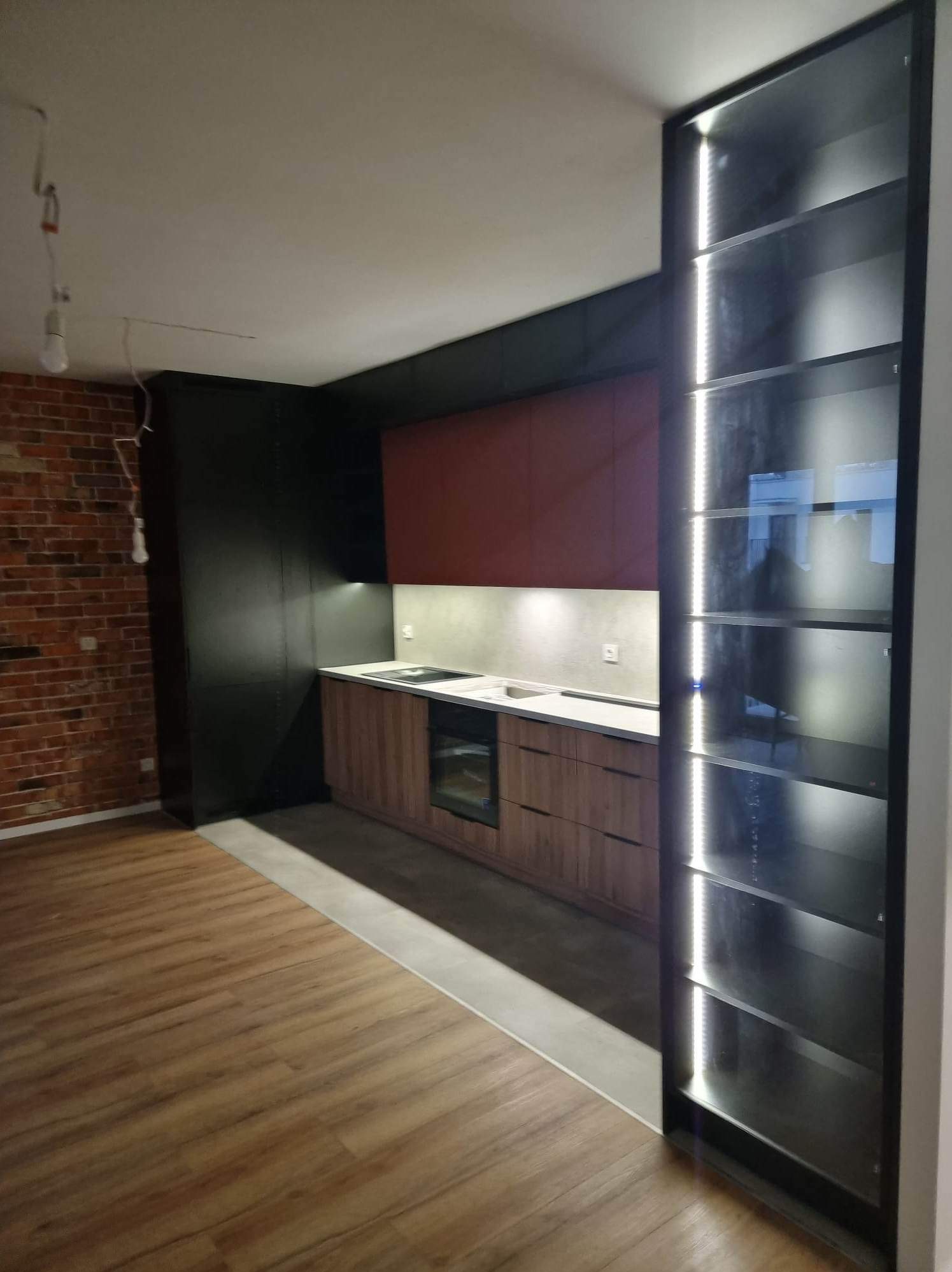 Nowoczesna kuchnia z drewnianymi frontami, czarnymi elementami i przeszkloną szafką z oświetleniem LED. Widoczna ceglana ściana i drewniana podłoga.