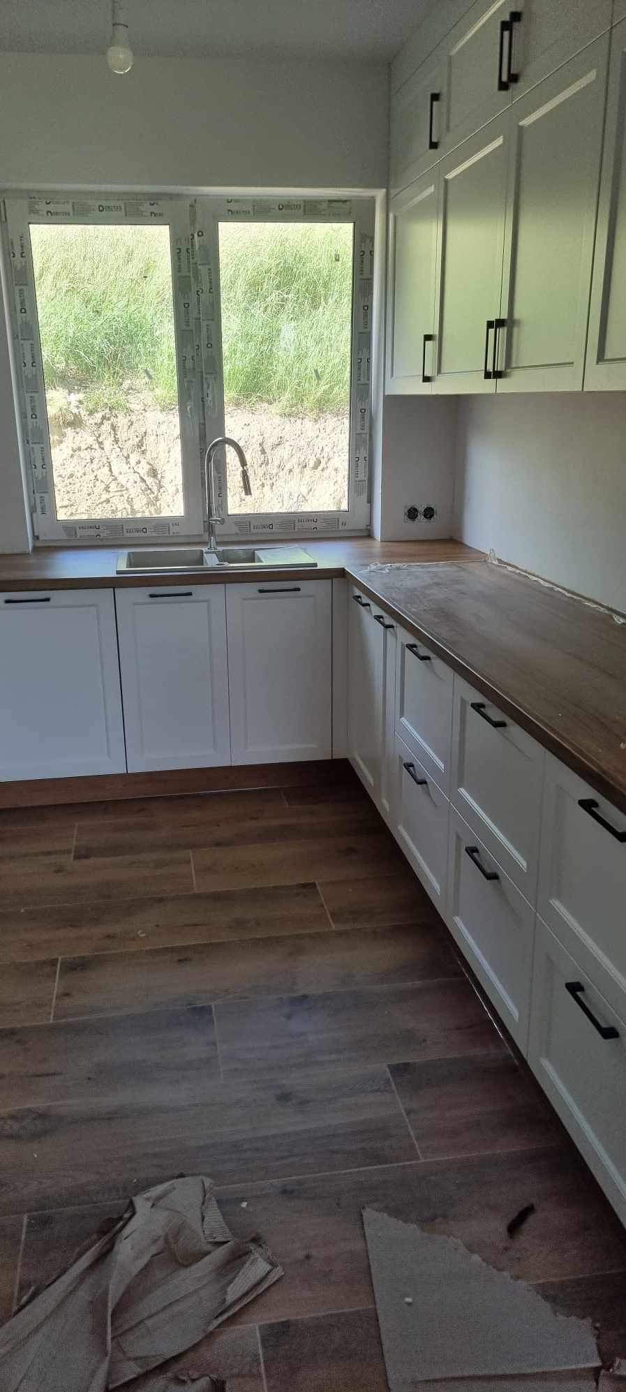 Jasna kuchnia z drewnianym blatem i czarnymi uchwytami, widok na okno z zielenią. Ułożone płytki podłogowe imitujące drewno. Fragment kartonu na podłodze.