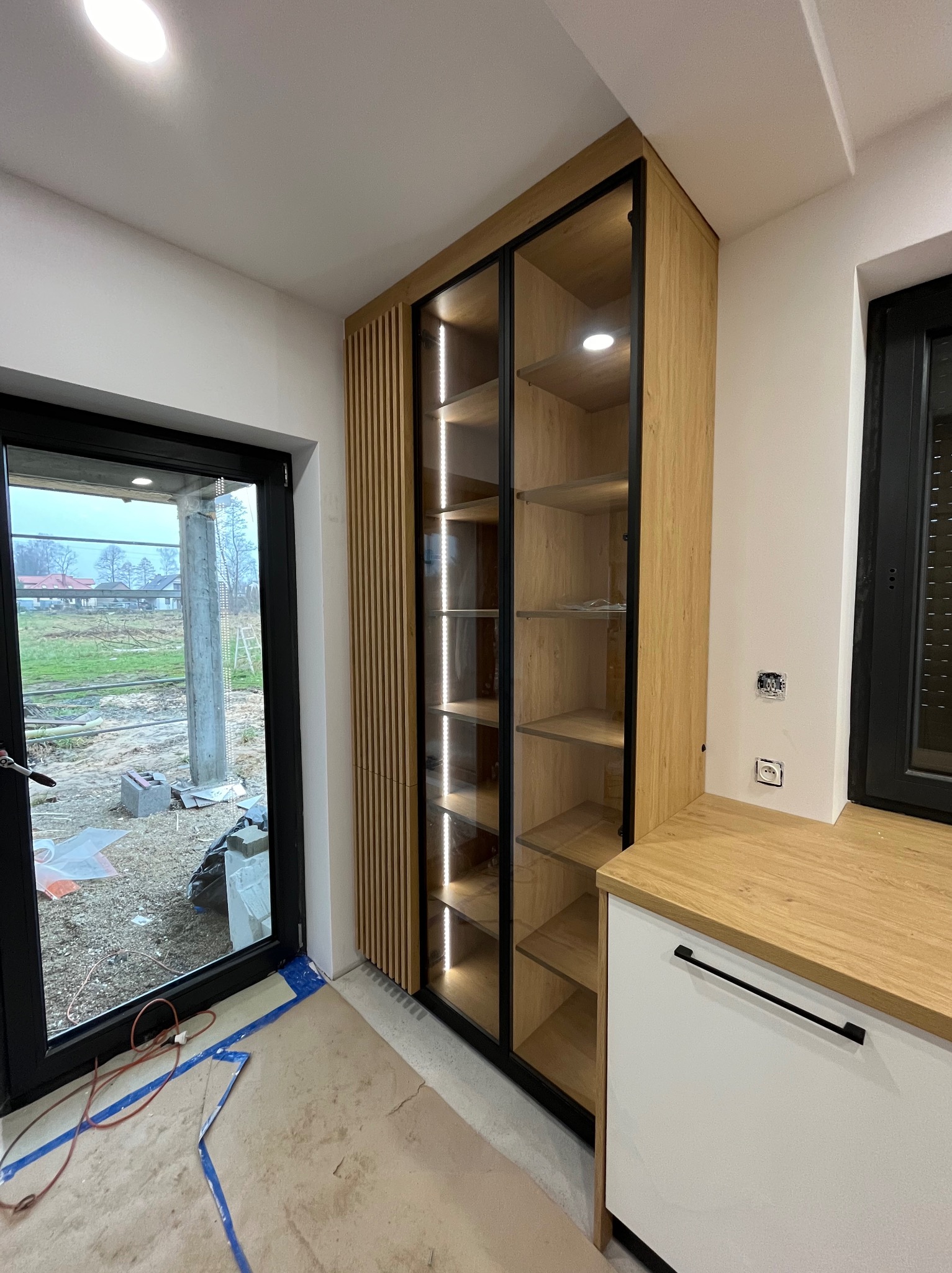 Nowoczesna szafa z przeszklonymi drzwiami i oświetleniem LED, zintegrowana z drewnianą zabudową. Widoczne elementy wykończenia wnętrza i okno z widokiem na zewnątrz.