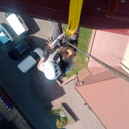 Pracownik w kasku, zabezpieczony liną, uszczelnia element dachu w Bytomiu, widok z góry na budynek z czerwonym dachem i zaparkowane samochody.