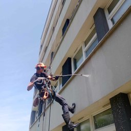 SkyRope Usługi Wysokościowe Paweł Krzystek - Pracownik w uprzęży alpinistycznej, z czarnymi gumowcami i pomarańczową czapką, myje elewację budynku wielorodzinnego przy użyciu lancy ciśnieniowej; woda spływa po ścianie, widoczne okna i ciemne...