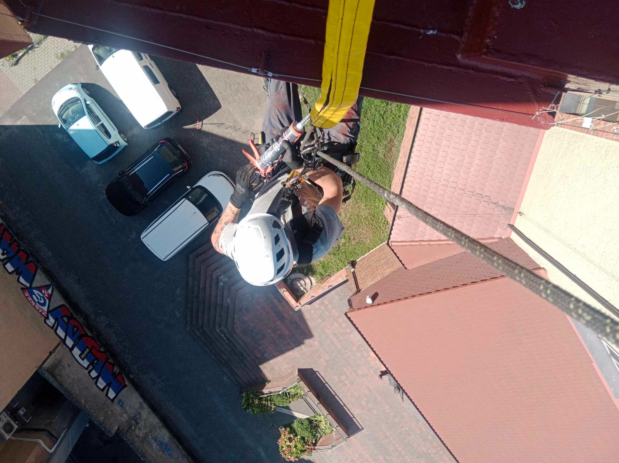 Pracownik w kasku, zabezpieczony liną, uszczelnia element dachu w Bytomiu, widok z góry na budynek z czerwonym dachem i zaparkowane samochody.