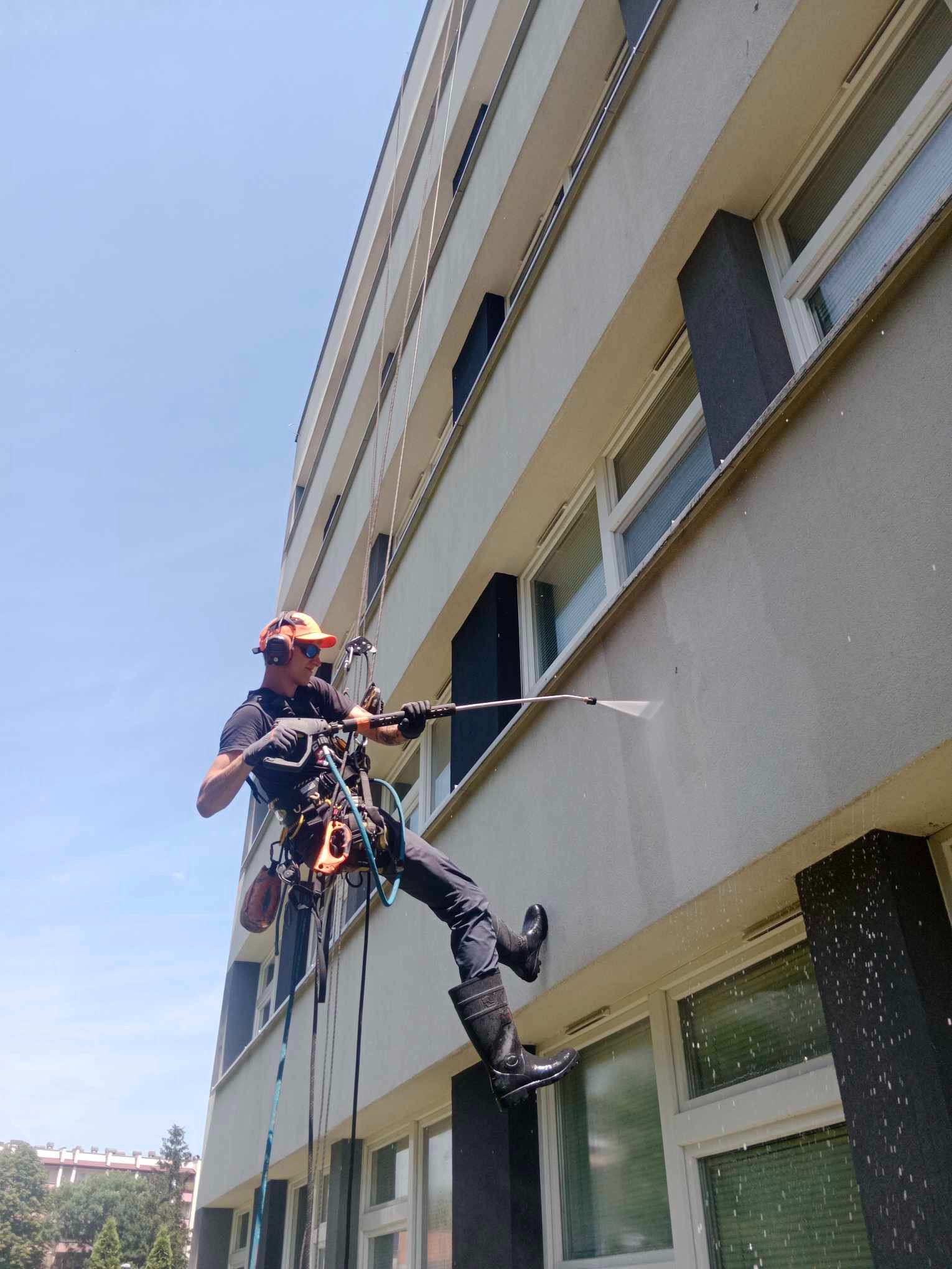 Pracownik w uprzęży alpinistycznej, z czarnymi gumowcami i pomarańczową czapką, myje elewację budynku wielorodzinnego przy użyciu lancy ciśnieniowej; woda spływa po ścianie, widoczne okna i ciemne...