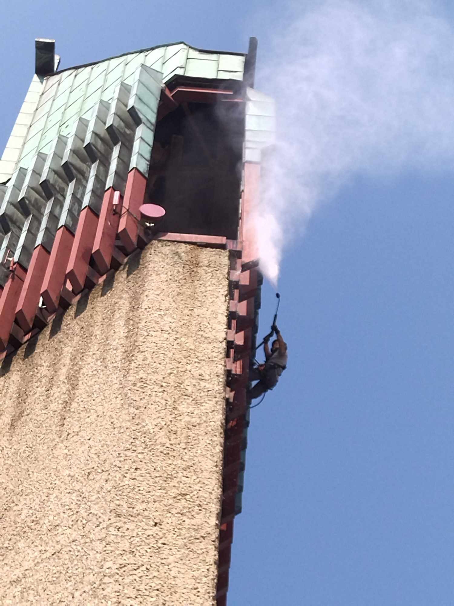 Pracownik na wysokości, zabezpieczony linami, myje elewację wieży wodą pod ciśnieniem, widoczny strumień wody i fragment dachu z zieloną dachówką.