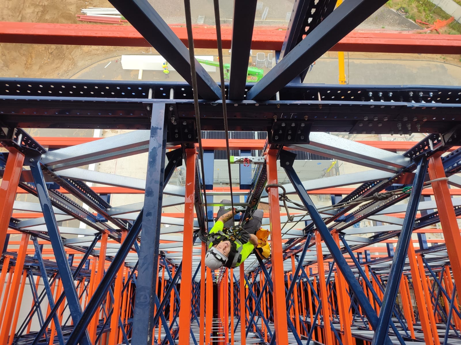 Pracownik w kasku i uprzęży, zawieszony na linach, pracuje na wysokości na metalowej konstrukcji pomalowanej na niebiesko i pomarańczowo, widok z dołu.