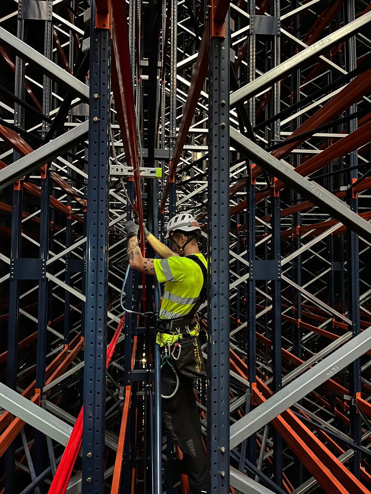 Pracownik w kasku i odzieży ochronnej, zabezpieczony linami, wykonuje prace na wysokości wewnątrz magazynu z regałami o konstrukcji metalowej w kolorach szarym, czerwonym i niebieskim.