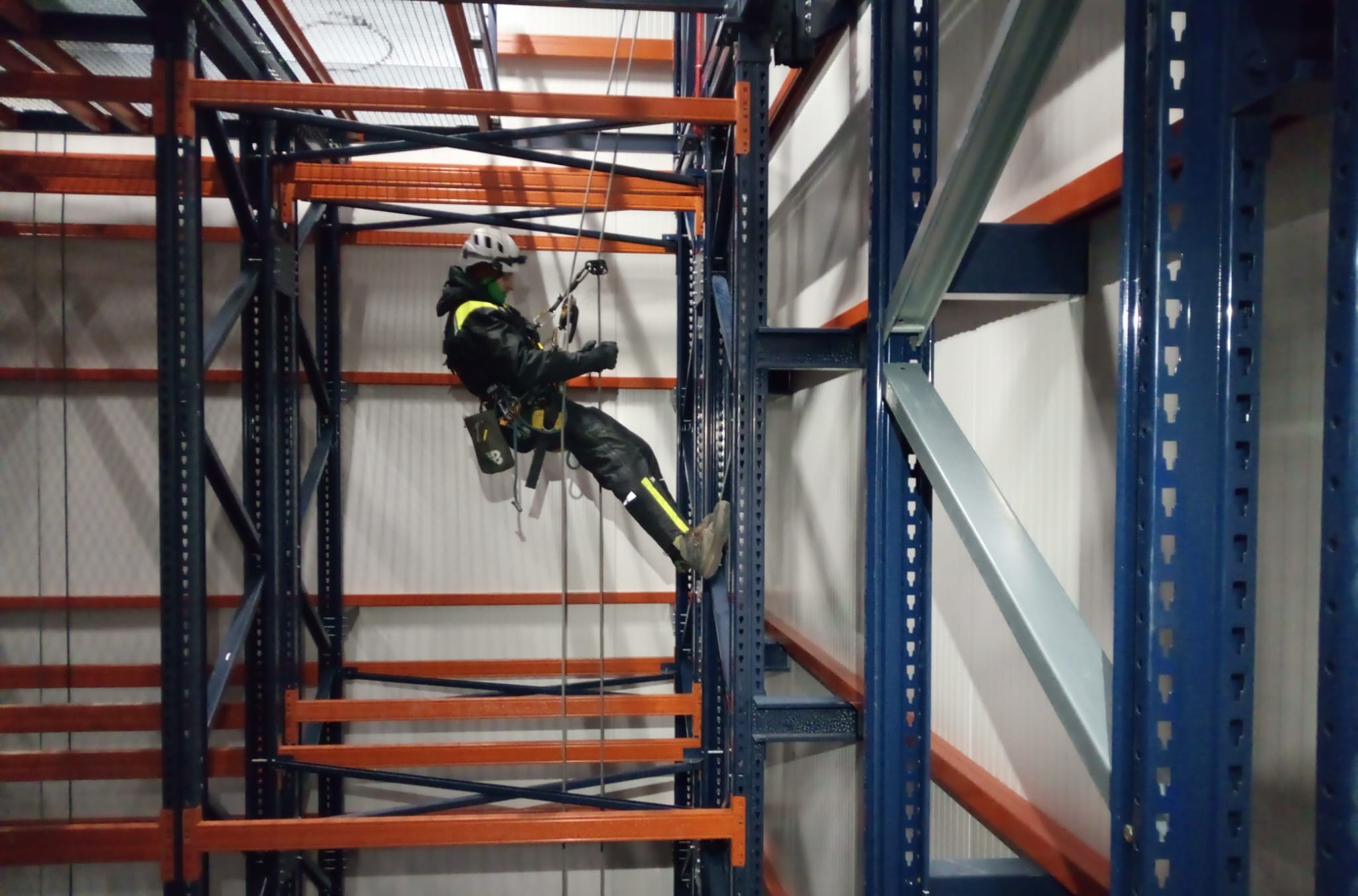 Pracownik w uprzęży, w kasku, na tle regałów magazynowych. Wykonywanie prac na wysokości w przestrzeni przemysłowej. Bezpieczeństwo i profesjonalizm.