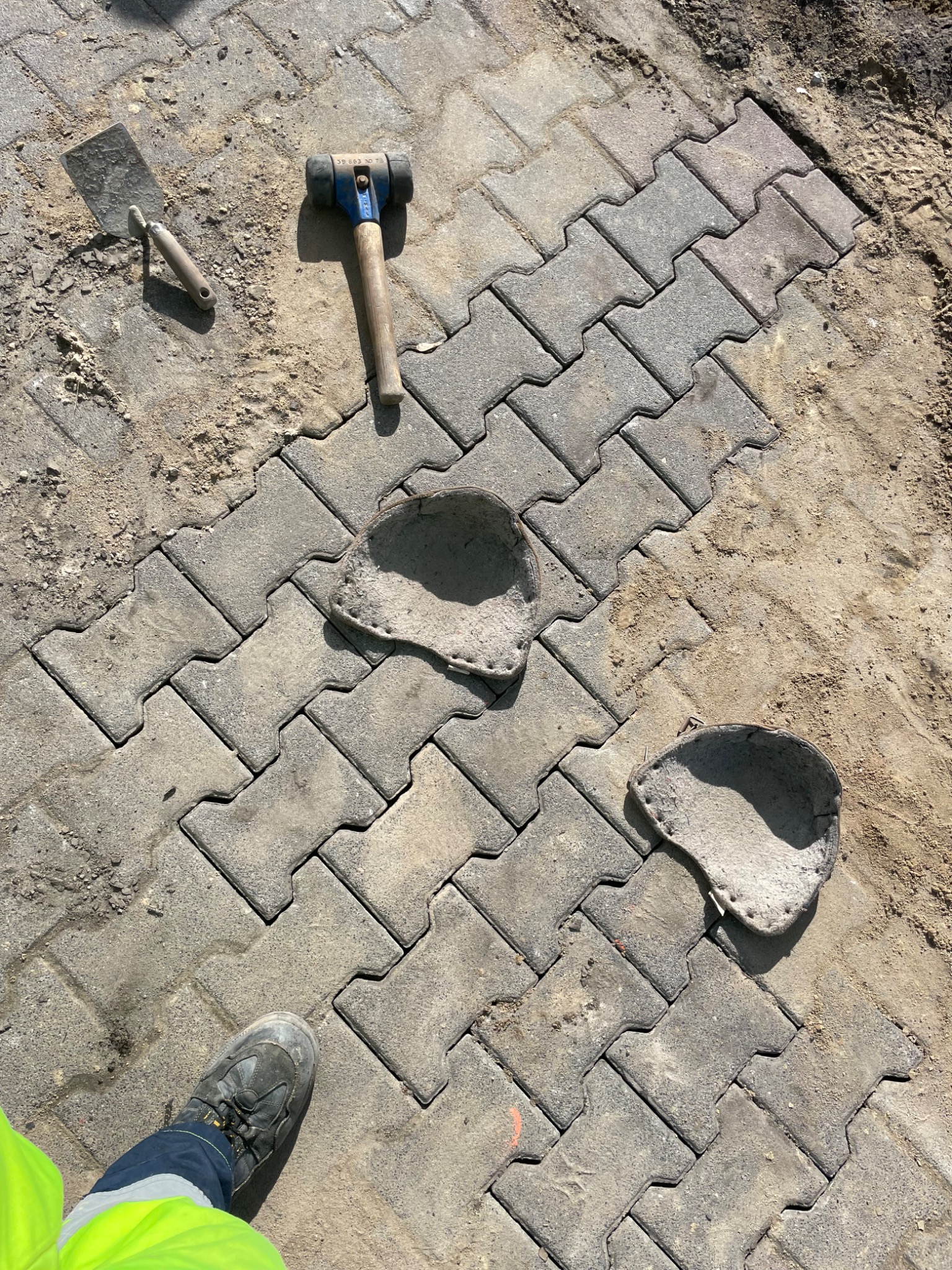Układanie szarej kostki brukowej typu 'I' z widocznymi narzędziami: gumowym młotkiem i kielnią, oraz wyciętymi miejscami na przyszłe nasadzenia roślin, obok stoi noga w roboczym bucie i odblaskowej...