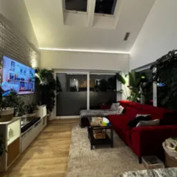 Nowoczesny salon z ceglaną ścianą, oświetleniem LED podsufitowym, czerwonym narożnikiem i licznymi roślinami doniczkowymi, oświetlony światłem z okien dachowych.