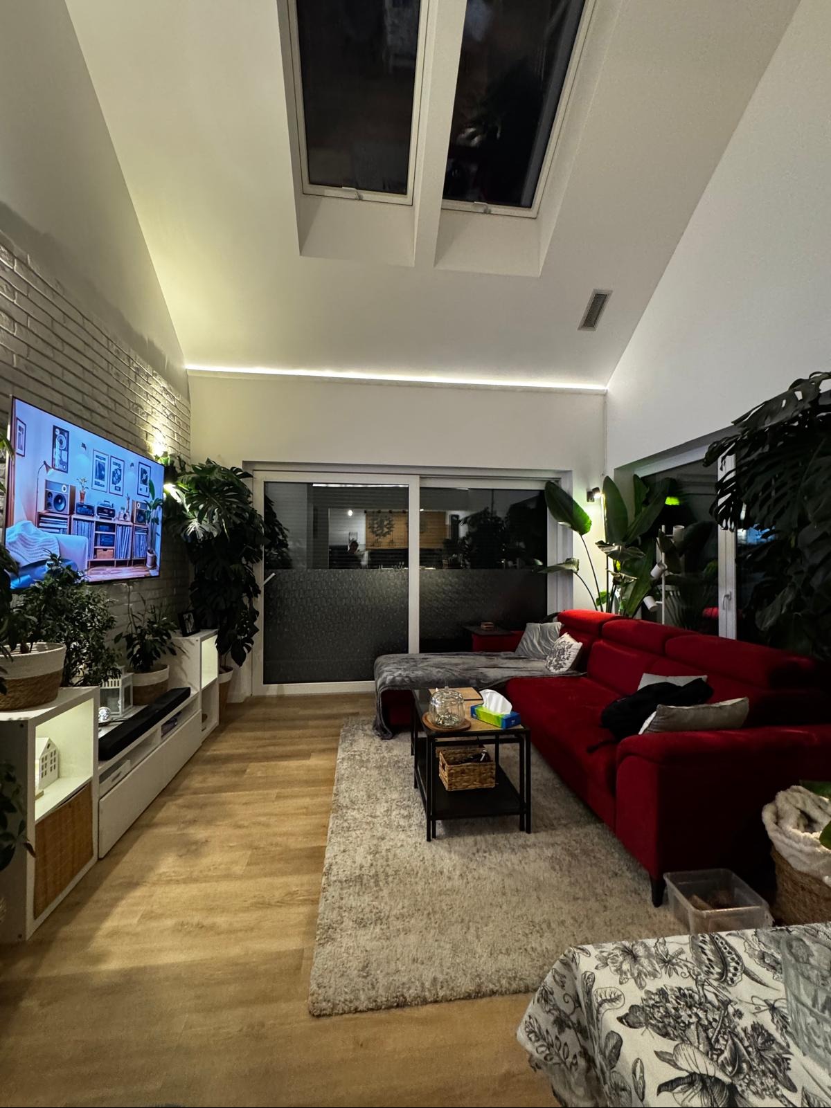 Nowoczesny salon z ceglaną ścianą, oświetleniem LED podsufitowym, czerwonym narożnikiem i licznymi roślinami doniczkowymi, oświetlony światłem z okien dachowych.