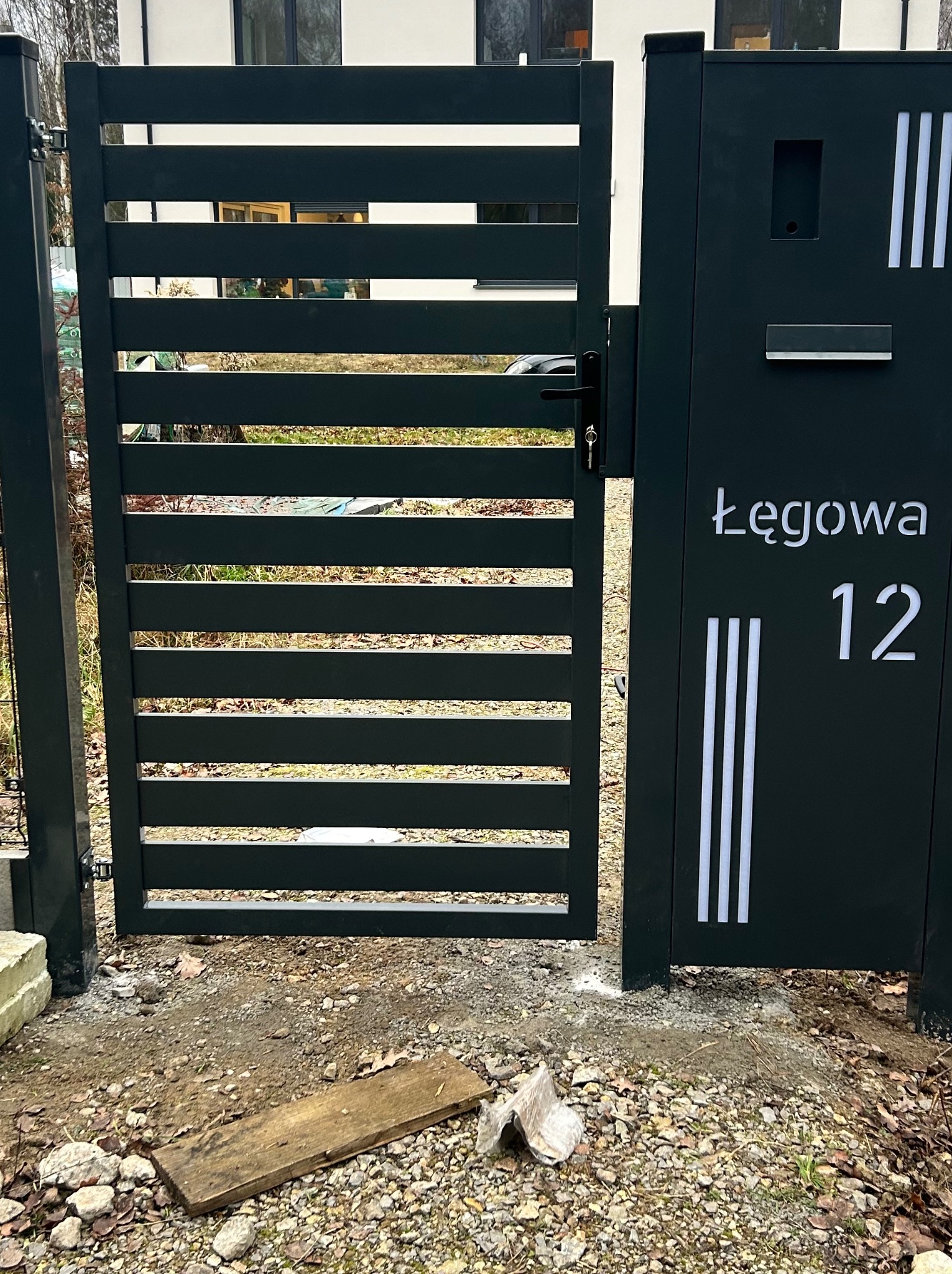 Nowoczesna, antracytowa furtka z poziomymi szczeblami i skrzynką na listy z adresem 'Łęgowa 12', na tle jasnego budynku. Widoczne detale wykończenia.
