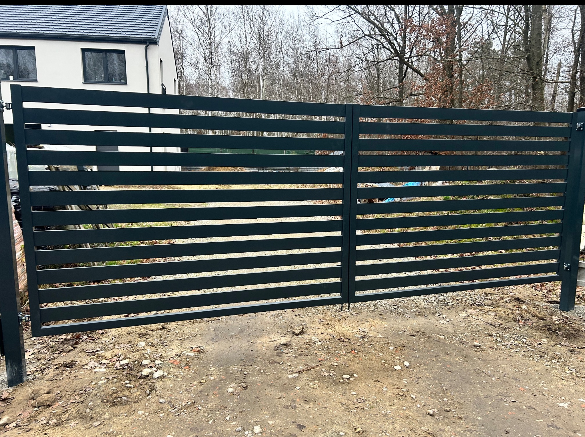 Nowoczesna, pozioma brama wjazdowa z metalowymi panelami w kolorze grafitowym, na tle domu i drzew. Minimalistyczny design, surowe wykończenie.