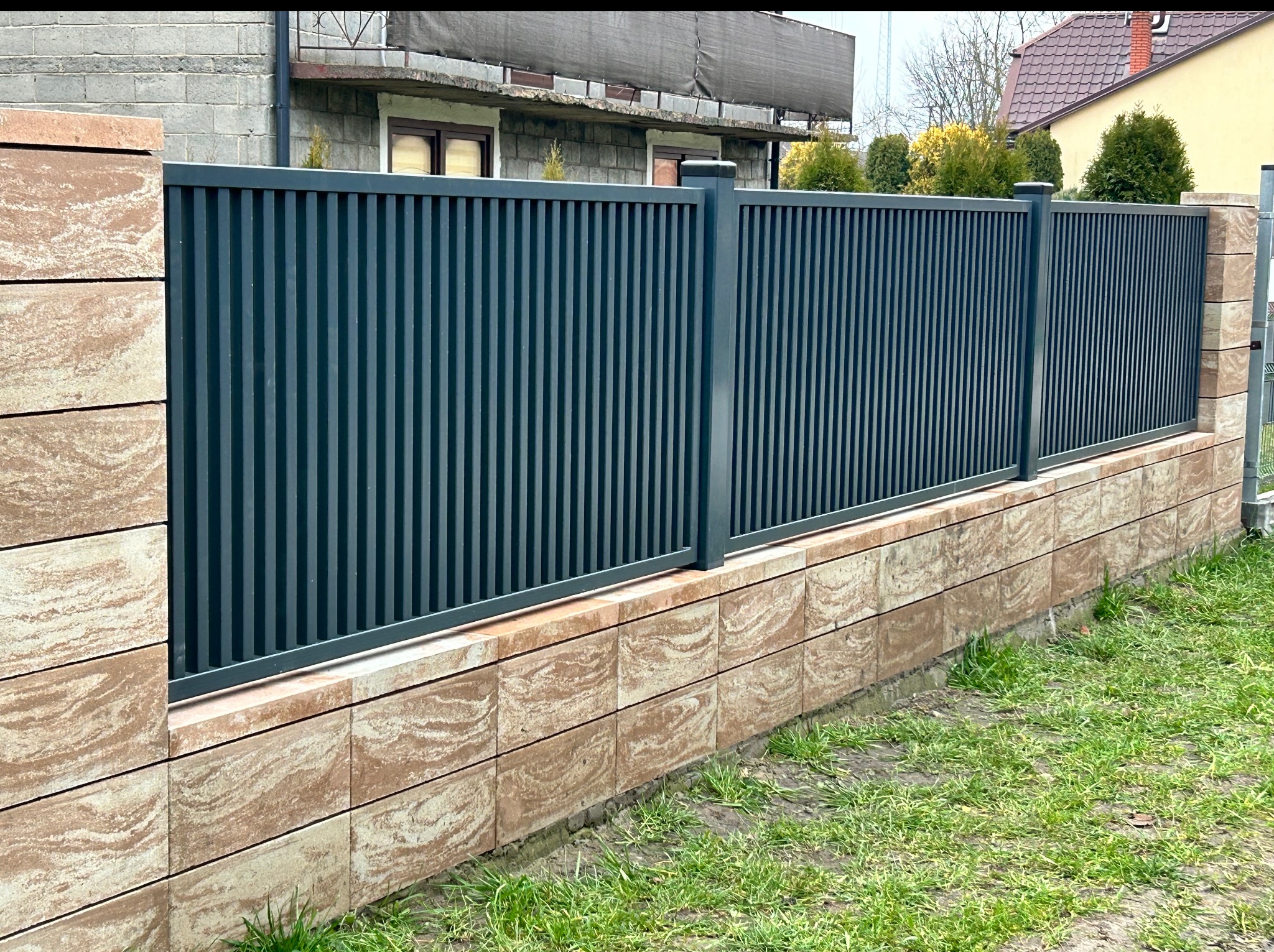 Nowoczesne, antracytowe ogrodzenie panelowe z podmurówką z dekoracyjnych bloczków w odcieniach beżu i brązu. Widok z perspektywy trawnika, dom w tle.