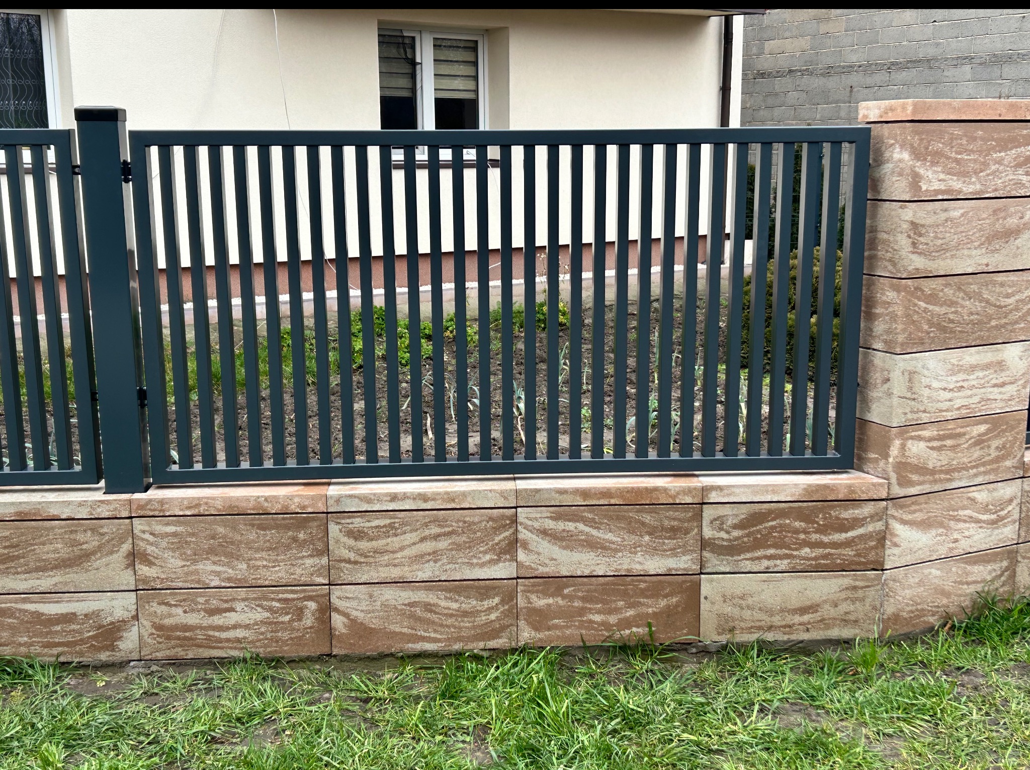 Nowoczesne ogrodzenie panelowe w kolorze grafitowym, osadzone na murku z dekoracyjnych bloczków w odcieniach beżu i brązu. Widok na posesję przez pionowe szczebelki.