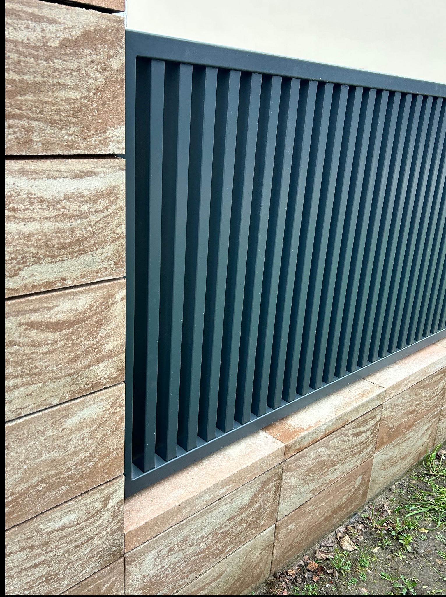 Nowoczesne, antracytowe ogrodzenie panelowe z pionowymi szczebelkami, osadzone na murku z trawertynu. Minimalistyczny design, widok z bliska, podkreślający detale.