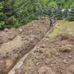M.K.Instal - Wykopany rów z ułożoną rurą kanalizacyjną i warstwą żwiru, pracownik kopie dalej, w tle drzewa iglaste.