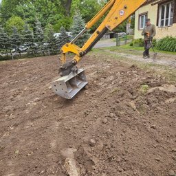 M.K.Instal - Żółta koparka z łyżką na nierównym terenie, w tle pracownik z łopatą i budynek mieszkalny.