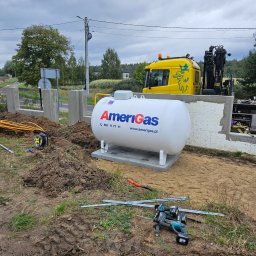 M.K.Instal - Biały zbiornik na gaz Amerigas na betonowej podstawie, żółty dźwig samochodowy w tle, narzędzia montażowe i rury na ziemi, częściowo ukończone ogrodzenie w tle.
