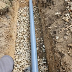 M.K.Instal - Szary rurociąg ułożony w wykopie z kamienistym dnem, widoczny fragment buta i nogawki pracownika.