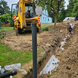 M.K.Instal - Wykopany rów z widoczną białą geowłókniną, czarna rura kanalizacyjna wystająca z ziemi, żółta koparko-ładowarka JCB w tle, pracownik z workiem, budynek mieszkalny i samochód dostawczy w oddali.