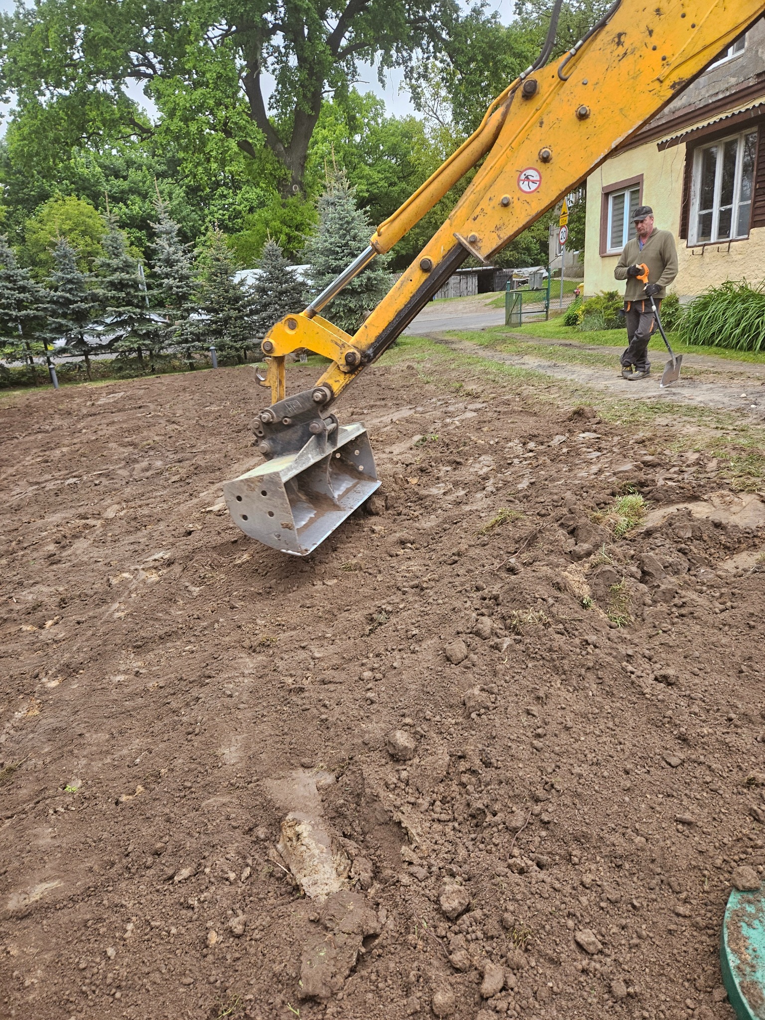 Żółta koparka z łyżką na nierównym terenie, w tle pracownik z łopatą i budynek mieszkalny.