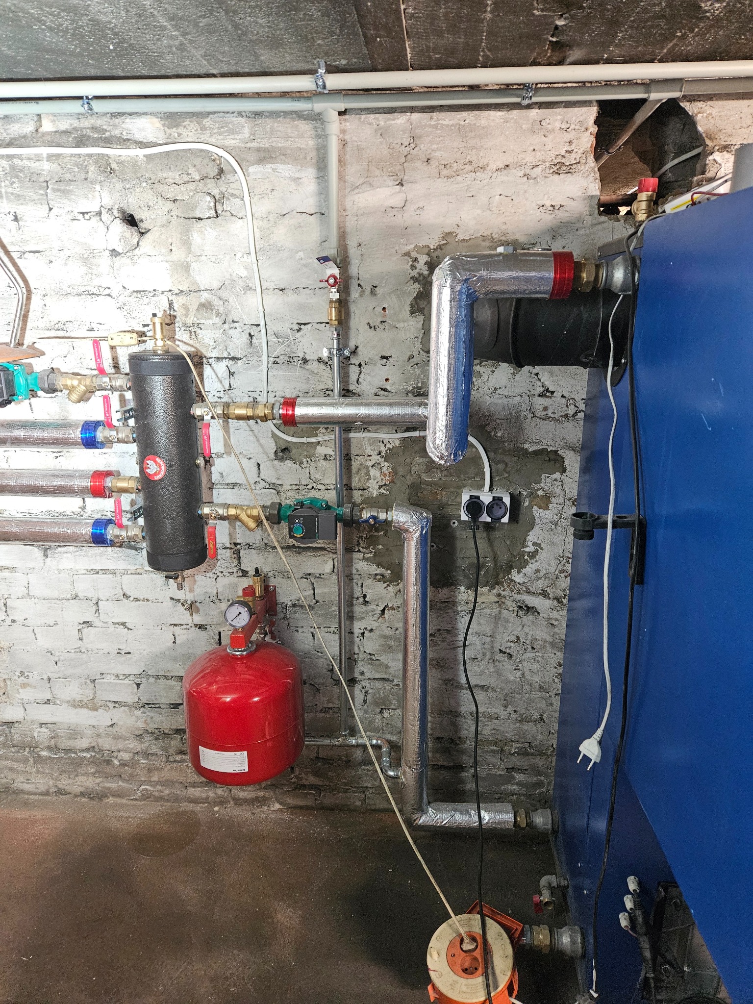 Instalacja grzewcza w kotłowni: widoczny czarny rozdzielacz z licznymi zaworami, czerwony zbiornik ciśnieniowy z manometrem, rury z izolacją aluminiową oraz fragment niebieskiego kotła na tle...