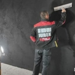 BARTŁOMIEJ JURKIEWICZ JUREK -BUD - Pracownik w firmowej kurtce nanosi szpachlą ciemną gładź na ścianę, widoczne oświetlenie robocze