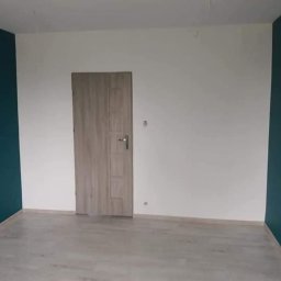 BARTŁOMIEJ JURKIEWICZ JUREK -BUD - Pomalowane na biało i turkusowo ściany w pokoju z szarą podłogą i drewnianymi drzwiami z srebrną klamką.