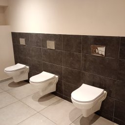 BARTŁOMIEJ JURKIEWICZ JUREK -BUD - Trzy podwieszane toalety z białymi deskami, zamontowane w rzędzie w wykończonej łazience z szarymi płytkami na ścianie i podłodze, widoczne przyciski spłukujące o różnym designie.