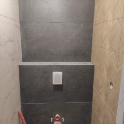 BARTŁOMIEJ JURKIEWICZ JUREK -BUD - Wykończona ściana w łazience z szarymi płytkami imitującymi kamień, obok płytki z wzorem jodełki, widoczny przycisk spłuczki i elementy instalacji sanitarnej