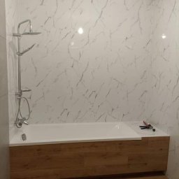 BARTŁOMIEJ JURKIEWICZ JUREK -BUD - Wykończona łazienka z wanną obudowaną panelami drewnopodobnymi, białe płytki imitujące marmur na ścianach, chromowana bateria prysznicowa, tubka silikonu i pistolet do silikonu leżące na obudowie...