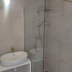 BARTŁOMIEJ JURKIEWICZ JUREK -BUD - Nowoczesna łazienka z owalną umywalką na białej szafce i kabiną prysznicową ze złotymi elementami, widoczny odpływ liniowy w podłodze.