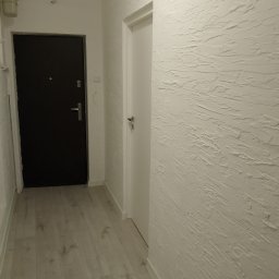 BARTŁOMIEJ JURKIEWICZ JUREK -BUD - Korytarz z dekoracyjną, białą ścianą o fakturze przypominającej gruboziarnisty tynk, ciemne drzwi wejściowe i białe drzwi wewnętrzne, wykończony panelami podłogowymi w odcieniach szarości.