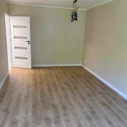 BARTŁOMIEJ JURKIEWICZ JUREK -BUD - Jasne panele podłogowe w pokoju z beżowymi ścianami, białe listwy przypodłogowe i drzwi z przeszkleniami. Nowoczesna lampa wisząca.