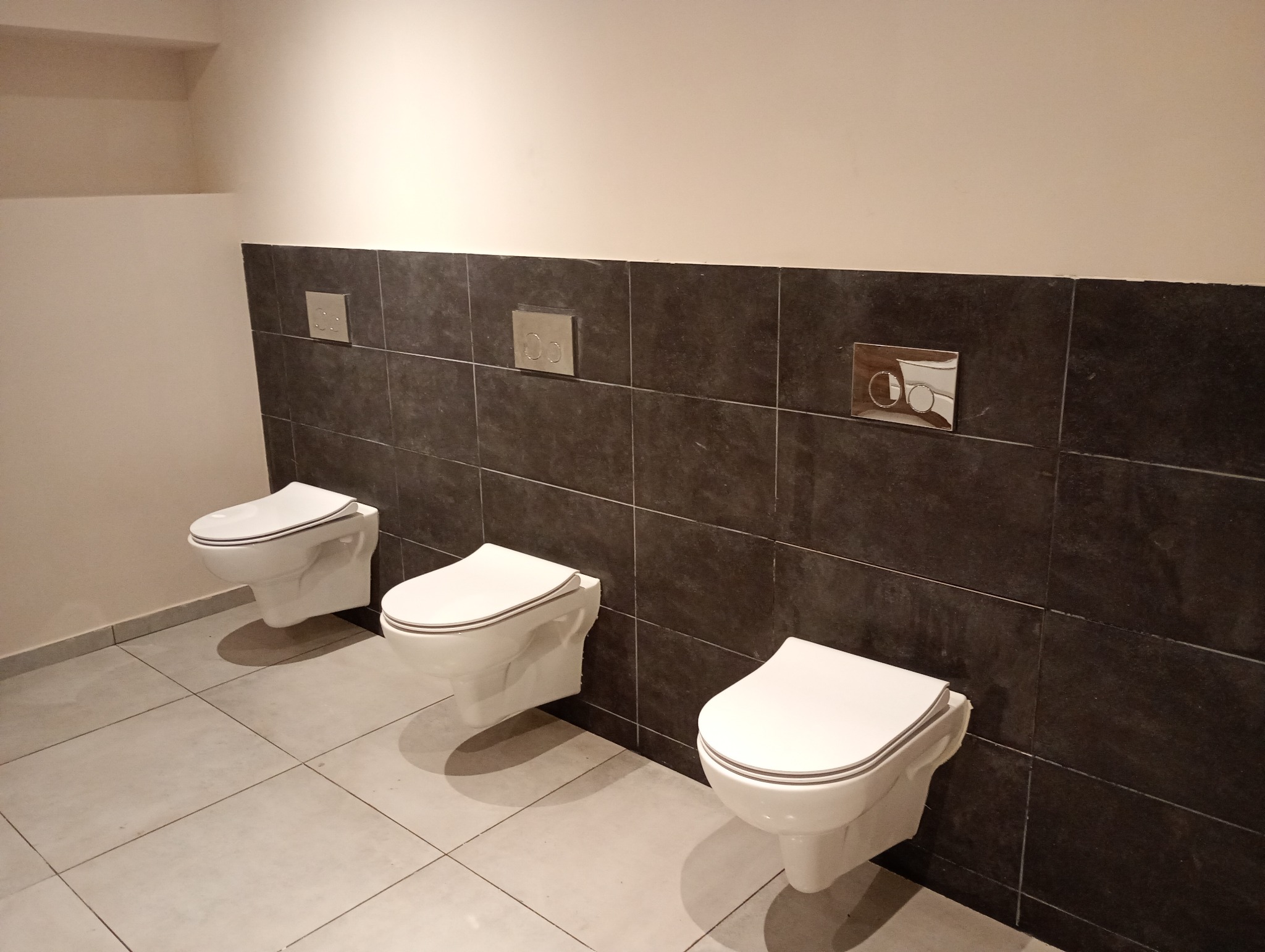 Trzy podwieszane toalety z białymi deskami, zamontowane w rzędzie w wykończonej łazience z szarymi płytkami na ścianie i podłodze, widoczne przyciski spłukujące o różnym designie.