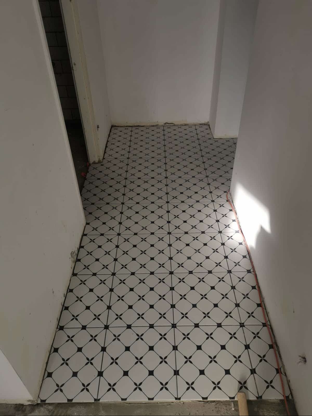 Ułożona podłoga w korytarzu z białych płytek z czarnym wzorem, widoczne niedociągnięcia przy ścianach i brak wykończenia przy progu.
