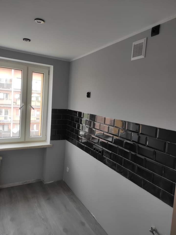 Szare wnętrze po remoncie z poziomym pasem czarnych, błyszczących płytek metra na ścianie, widok na okno i szarą podłogę.