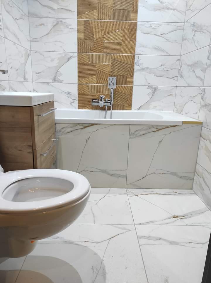 Nowoczesna łazienka z biało-złotymi płytkami imitującymi marmur, akcentem z drewnopodobnych płytek nad wanną, białą wanną z zabudową, umywalką z szafką w kolorze drewna i wiszącą toaletą.