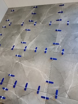 Układanie płytek ceramicznych na podłodze z użyciem niebieskich klipsów poziomujących i czarnej nici krzyżowej, widoczne szare płytki imitujące marmur.