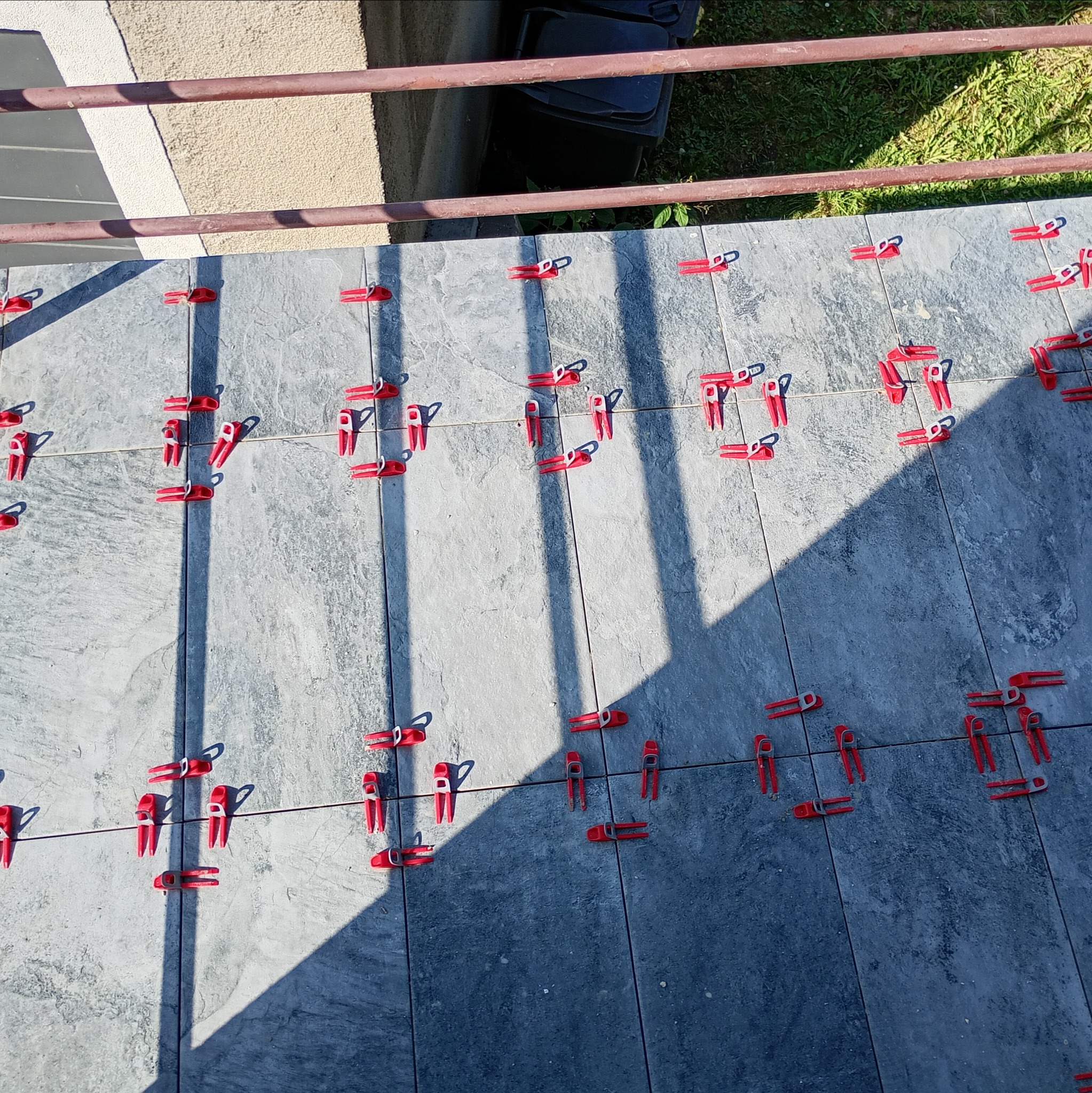 Szare płytki podłogowe układane z użyciem czerwonych klipsów poziomujących, widoczne cienie, fragment balkonu z brązową barierką i trawa w tle.